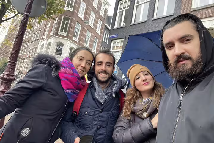 Four friends smiling together on a rainy Amsterdam street with historic canal houses in the background