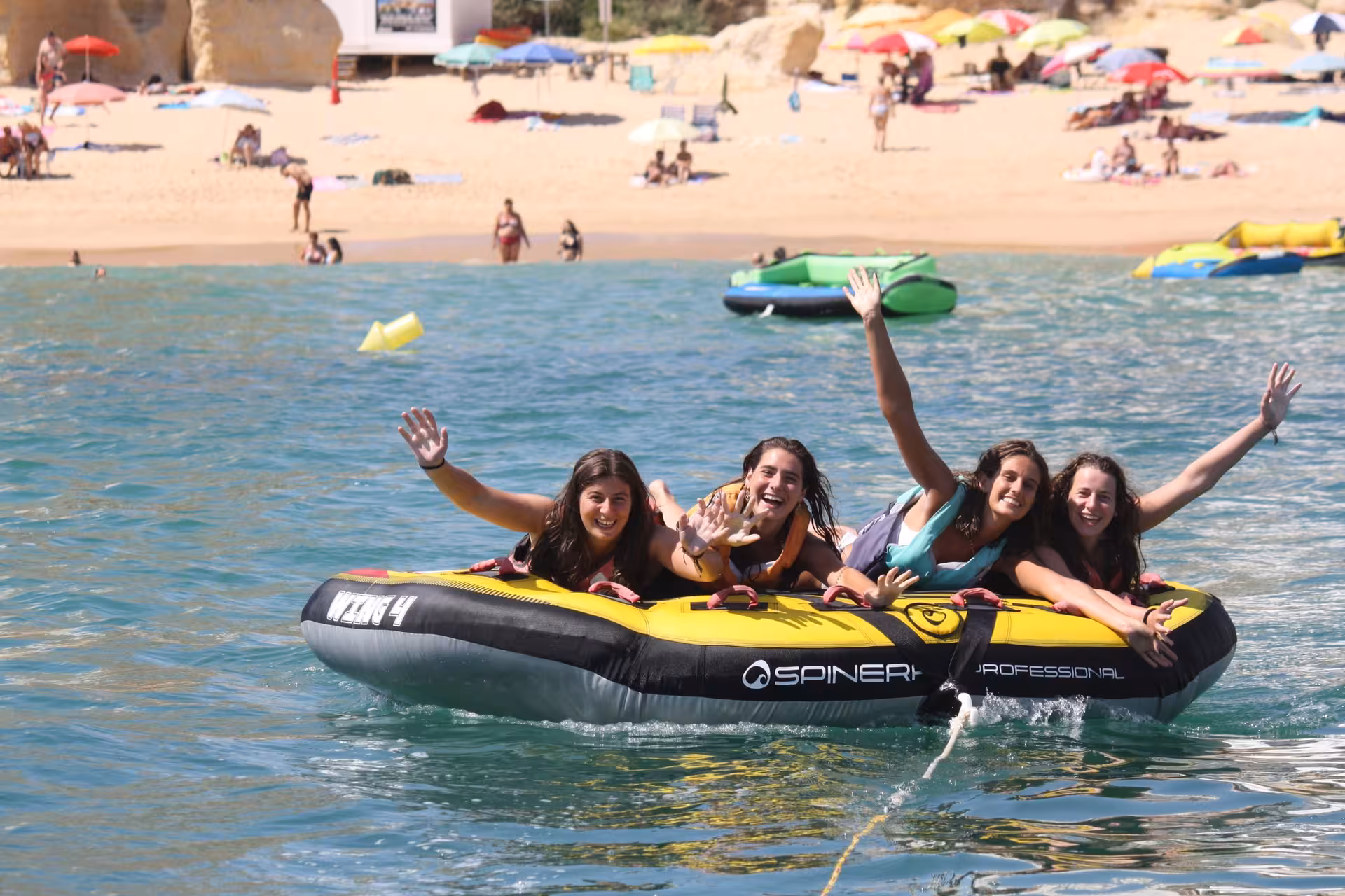 Four cheerful girls waving hands on a yellow Spinera inflatable tube ride near a vibrant Algarve beach shoreline