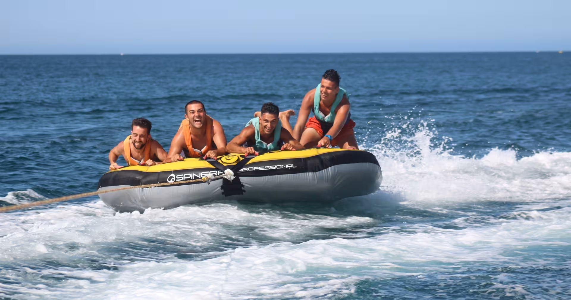Four friends laughing and riding the Crazy Cookie inflatable tube towed across thrilling ocean waves on a sunny day.
