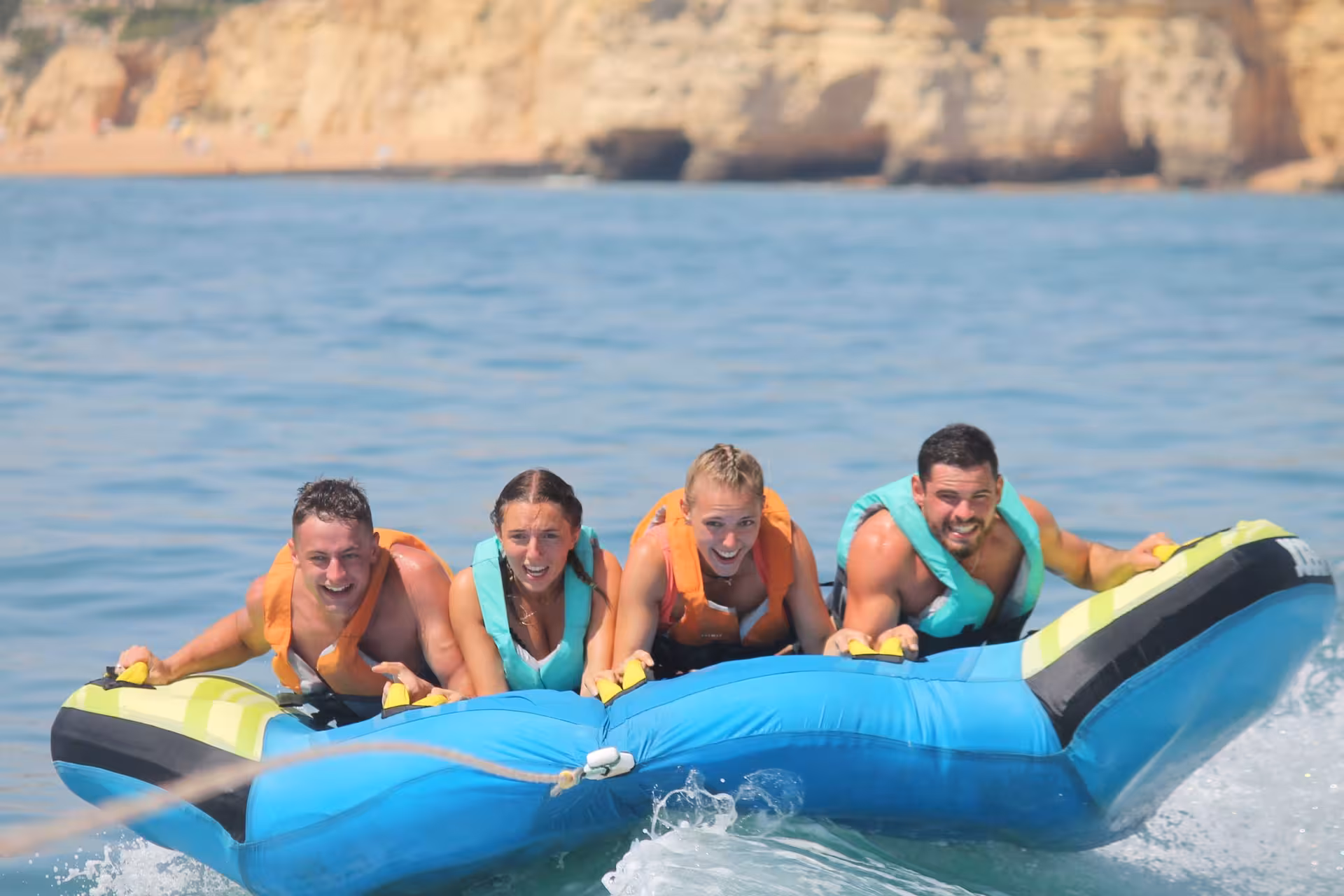 Four smiling friends in life jackets riding a blue inflatable Crazy Cookie tube towed across sparkling ocean waters