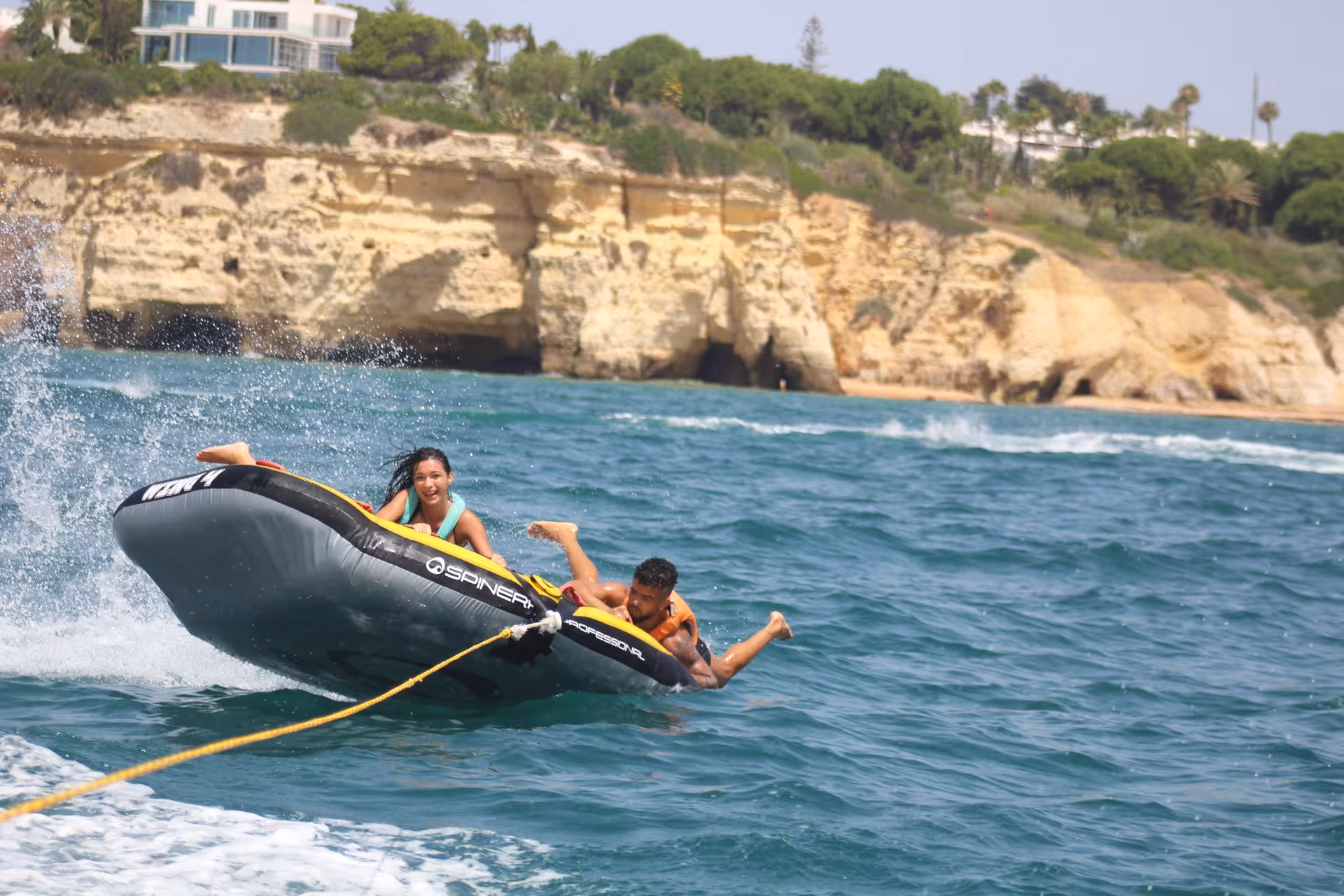 Two riders on a Spinera inflatable tube tilting sideways near Algarve cliffs during a thrilling Crazy Cookie watersports tubing experience