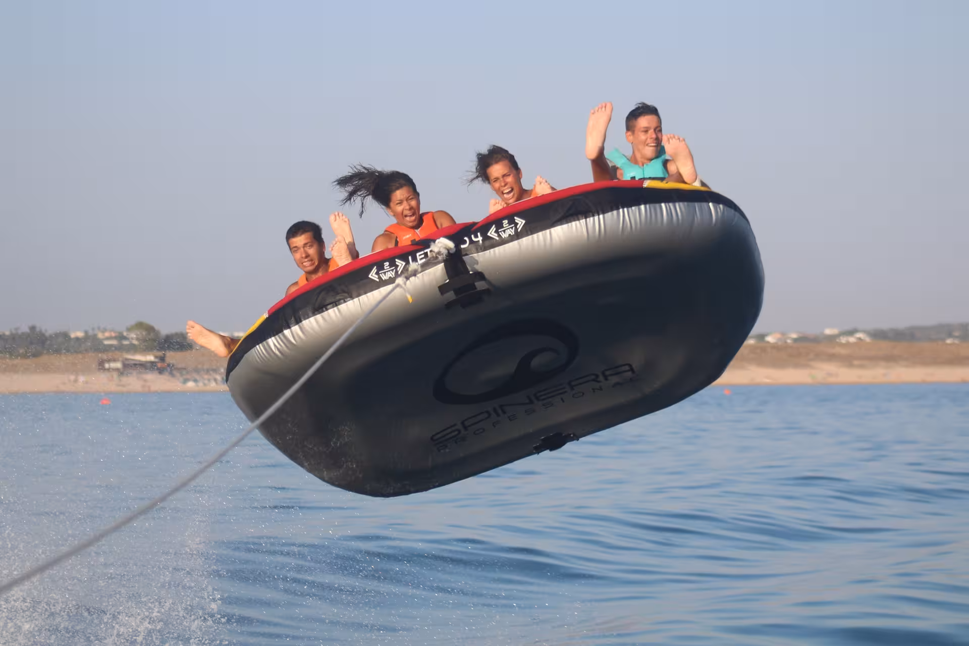 Four excited riders soaring airborne on a Crazy Sofa inflatable tube towed by a speedboat over sparkling ocean