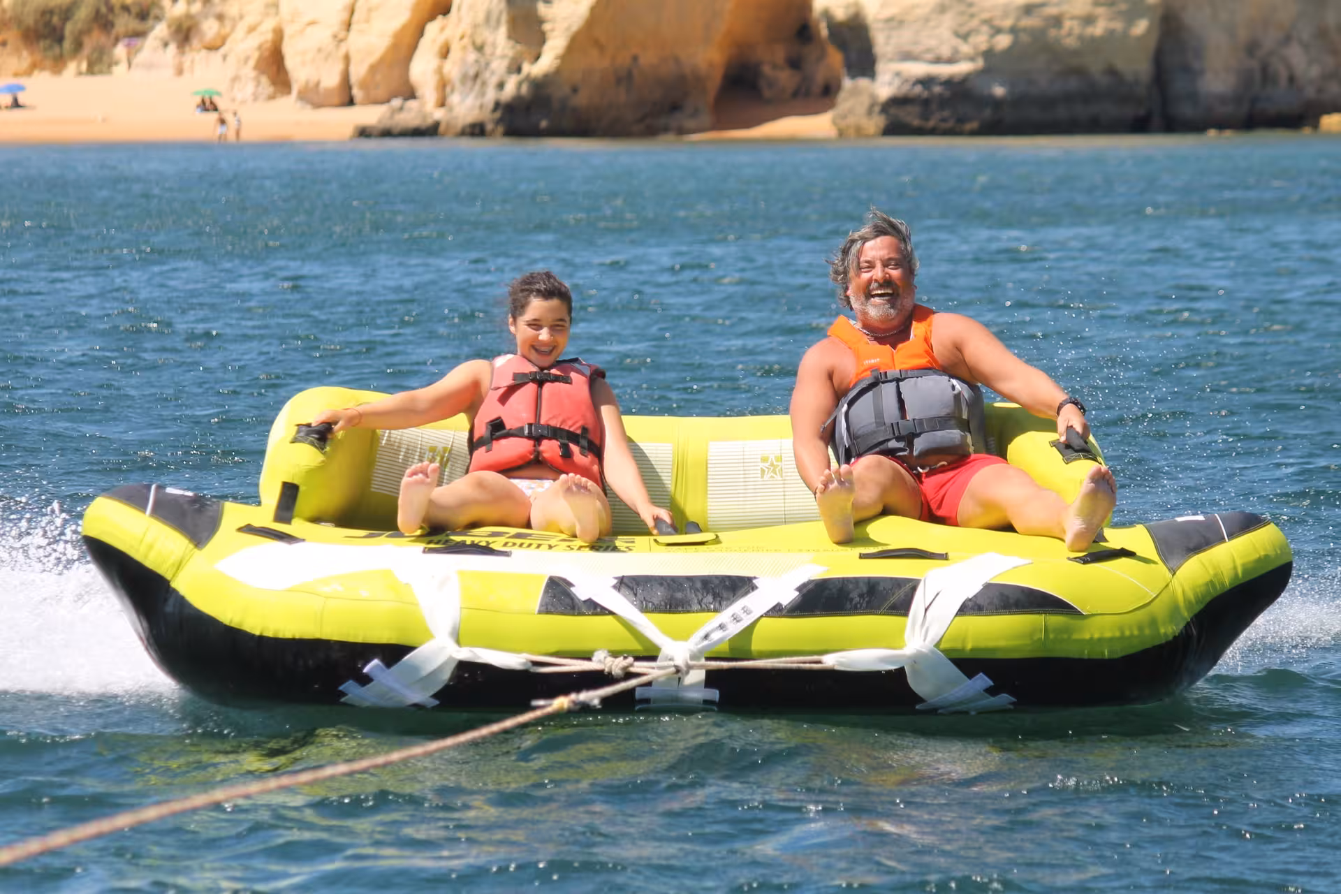 Father and child laughing on a Crazy Sofa inflatable water tube ride along the Algarve coastline