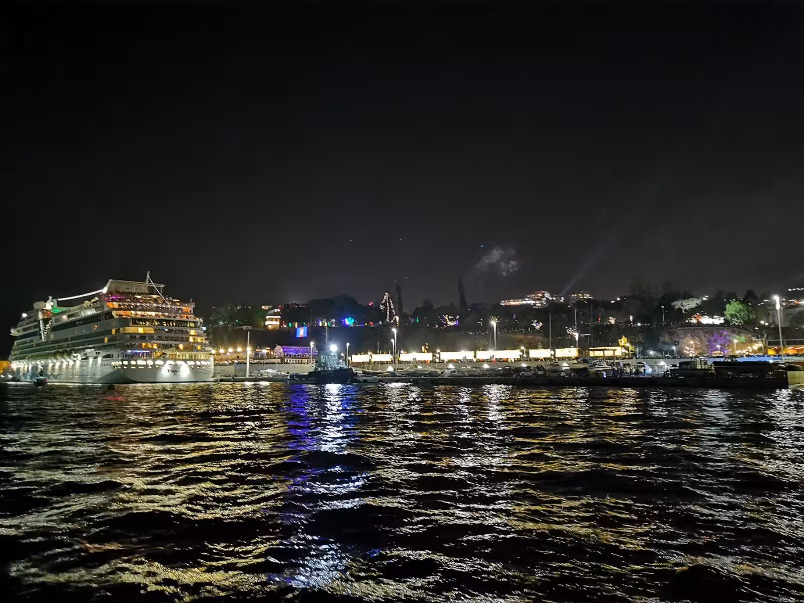 Cruise ship docked at a brightly lit harbor during New Year's Eve, with reflections shimmering on the water.