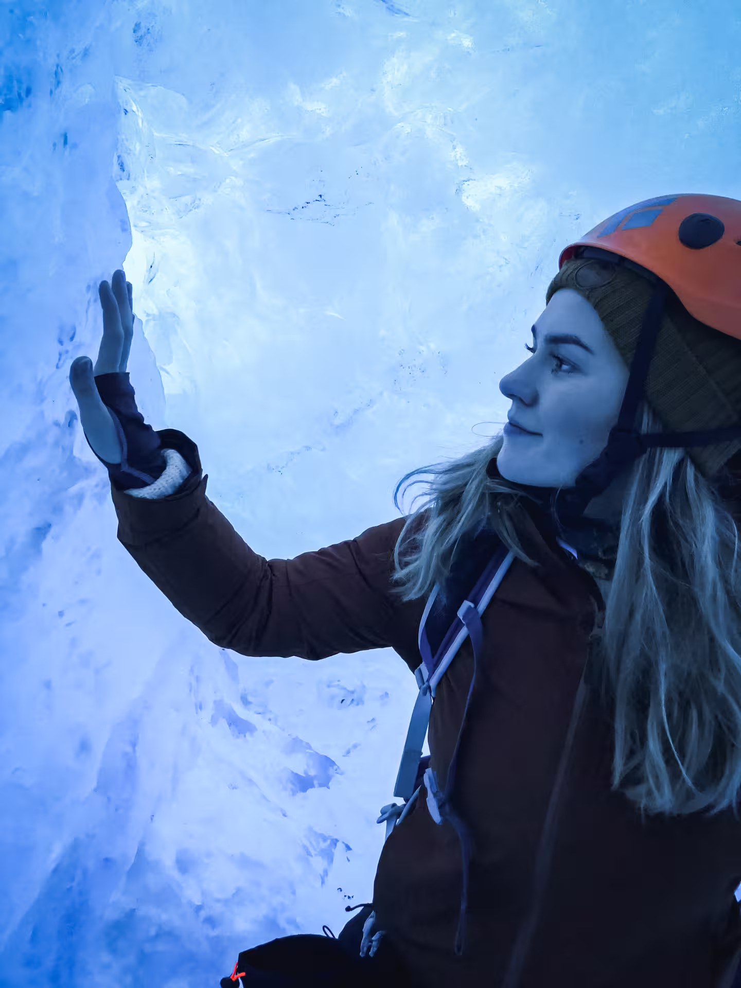 Woman in orange helmet touching glowing blue ice wall inside Vatnajokull glacier ice cave