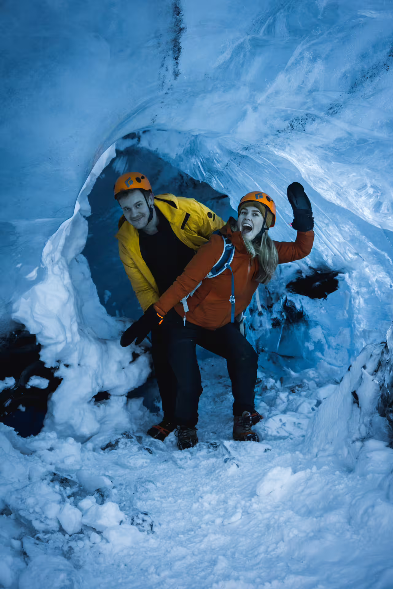 Two explorers in orange helmets and colorful jackets posing inside a blue ice cave at Vatnajokull Glacier
