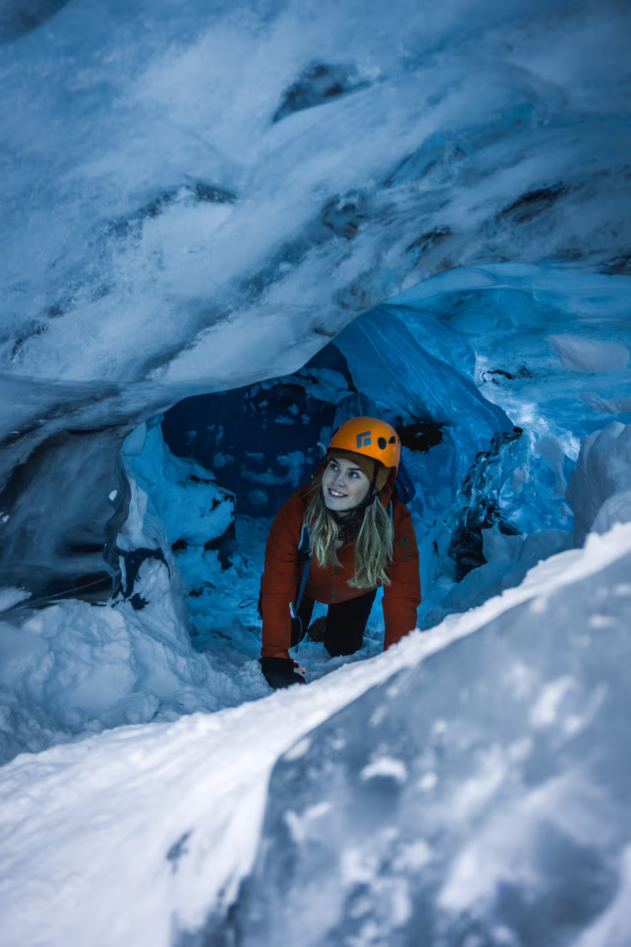 Smiling woman in orange helmet and jacket emerging from a blue ice cave at Vatnajokull Glacier