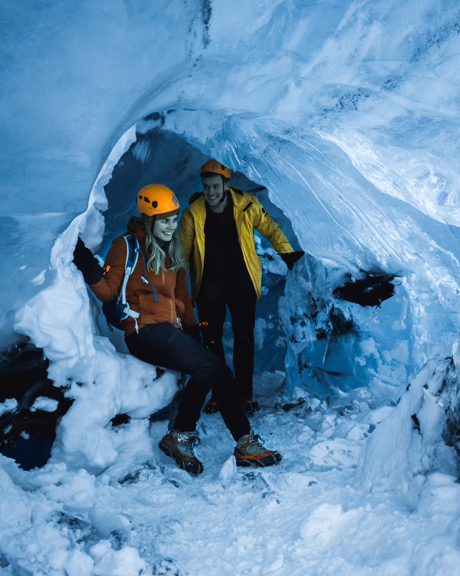 Two smiling guides in orange helmets exploring the blue ice cave tunnel at Vatnajokull Glacier