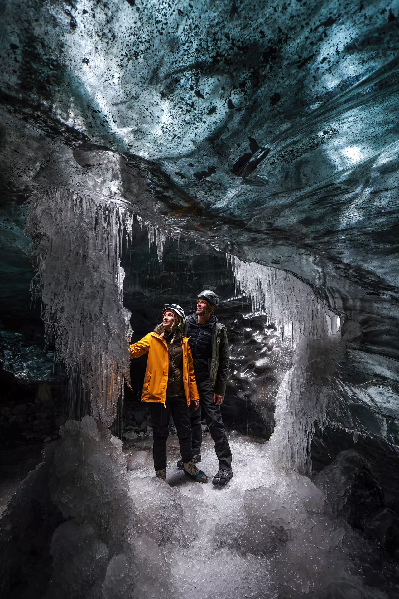 A couple in helmets and winter gear stands beneath dramatic icicles in a crystal ice cave on Vatnajokull Glacier Iceland.