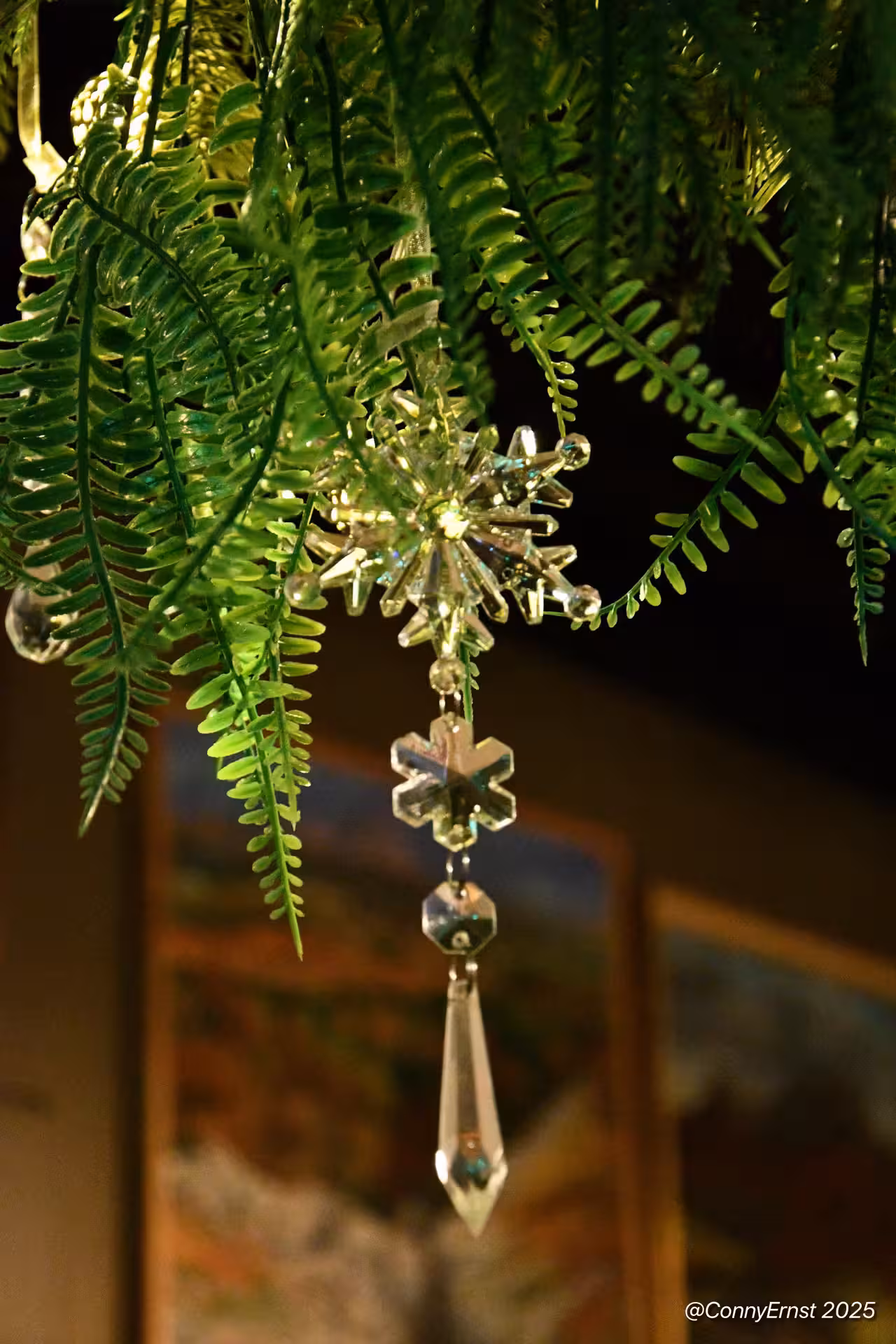 Crystal snowflake ornament hanging from lush green fern, sparkling with light in an indoor setting