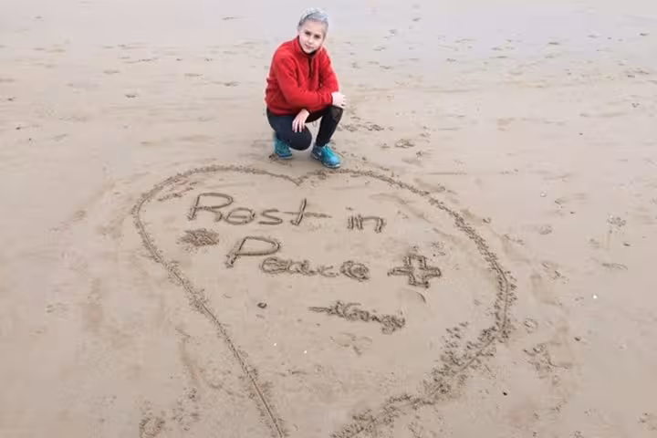 Person crouching beside heart drawn in sand with Rest in Peace message on D-Day beach in Normandy