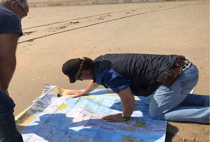Tour guide kneeling on beach examining large D-Day map with visitor during Normandy tour