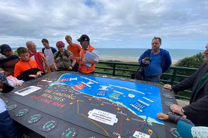 Tour group gathered around D-Day landing beaches map at Normandy overlook with ocean in background