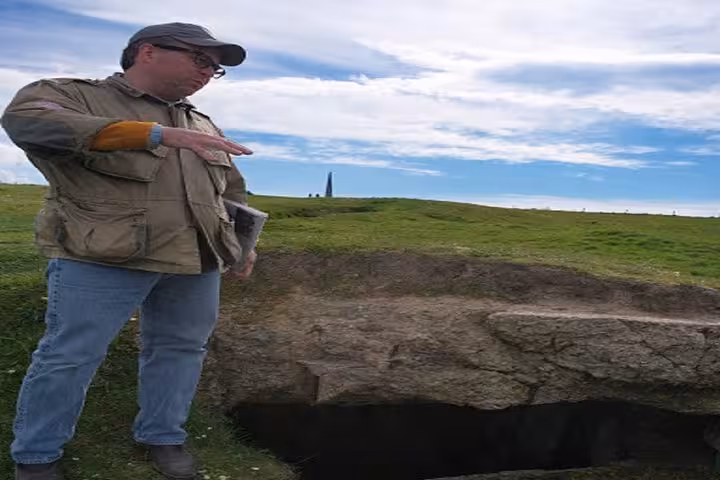 Tour guide pointing at D-Day bunker excavation on Normandy battlefield with monument in background