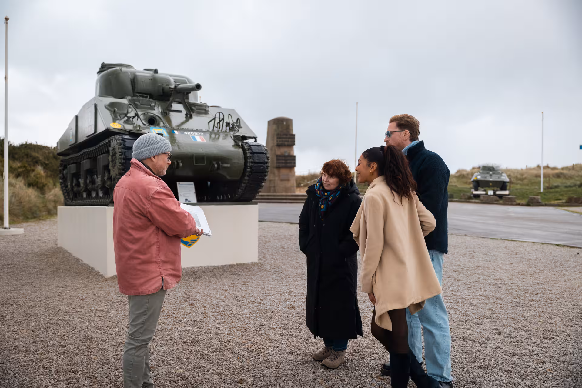 Tour guide explaining Sherman tank display to tourists at D-Day landing beach memorial in Normandy