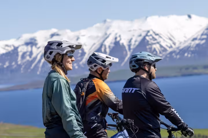 Three mountain bikers wearing helmets pause with e-bikes overlooking snowy mountains and a lake in Dalvik, Iceland