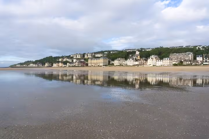 Deauville beach shoreline reflecting elegant Belle Époque buildings and green hillside under cloudy sky