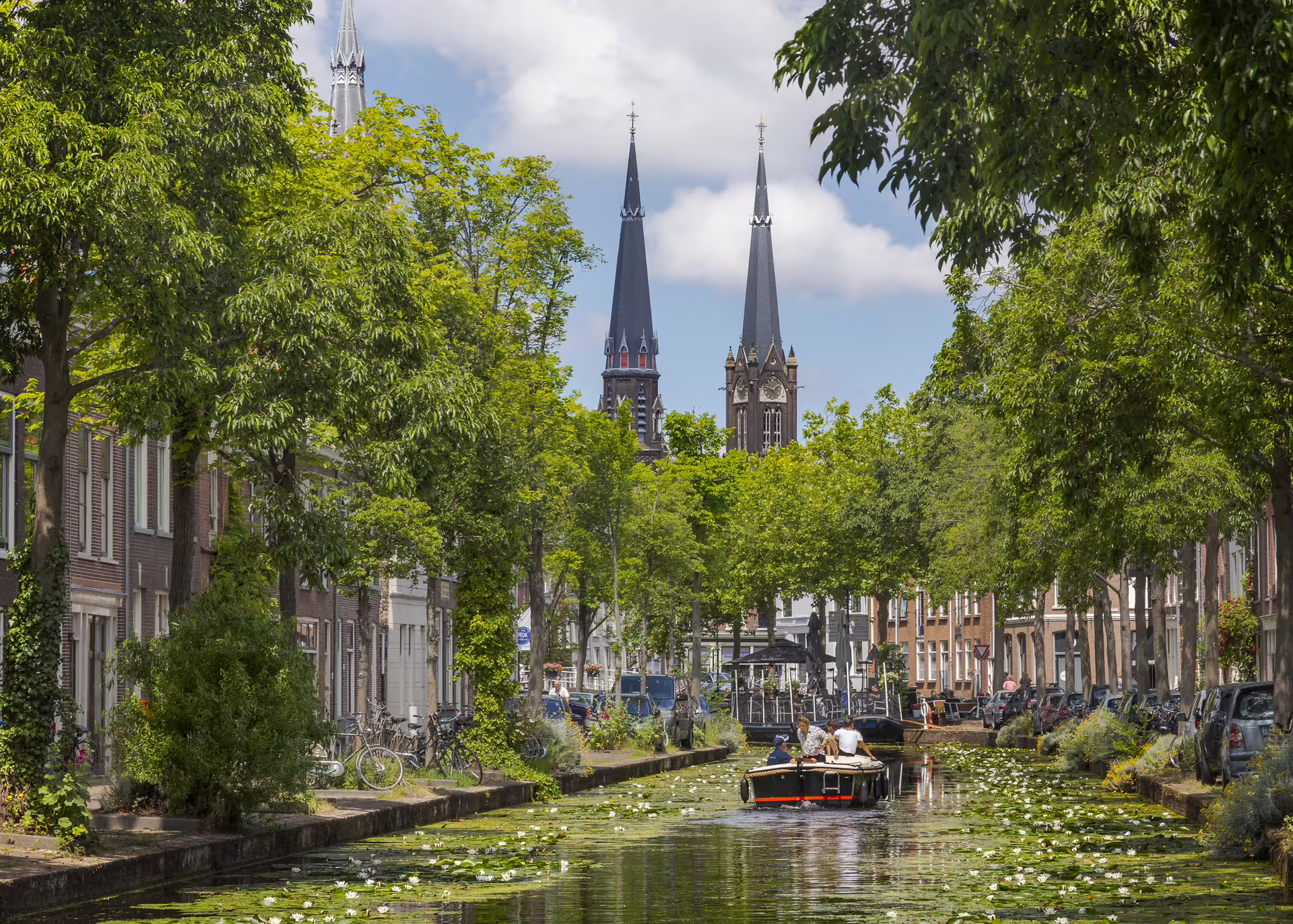 Delft canal with lily pads and boat, framed by trees and church spires on 1-day audioguide walk