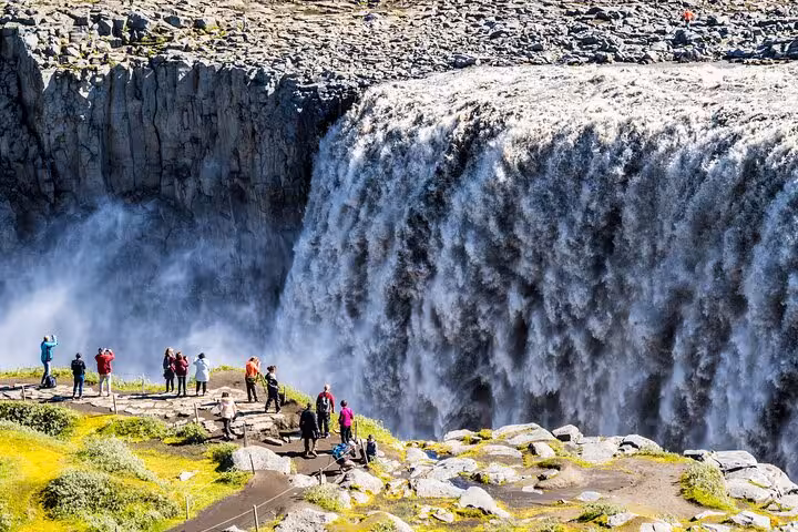 Visitors capture the majestic flow of Dettifoss waterfall, surrounded by lush greenery and rocky cliffs.