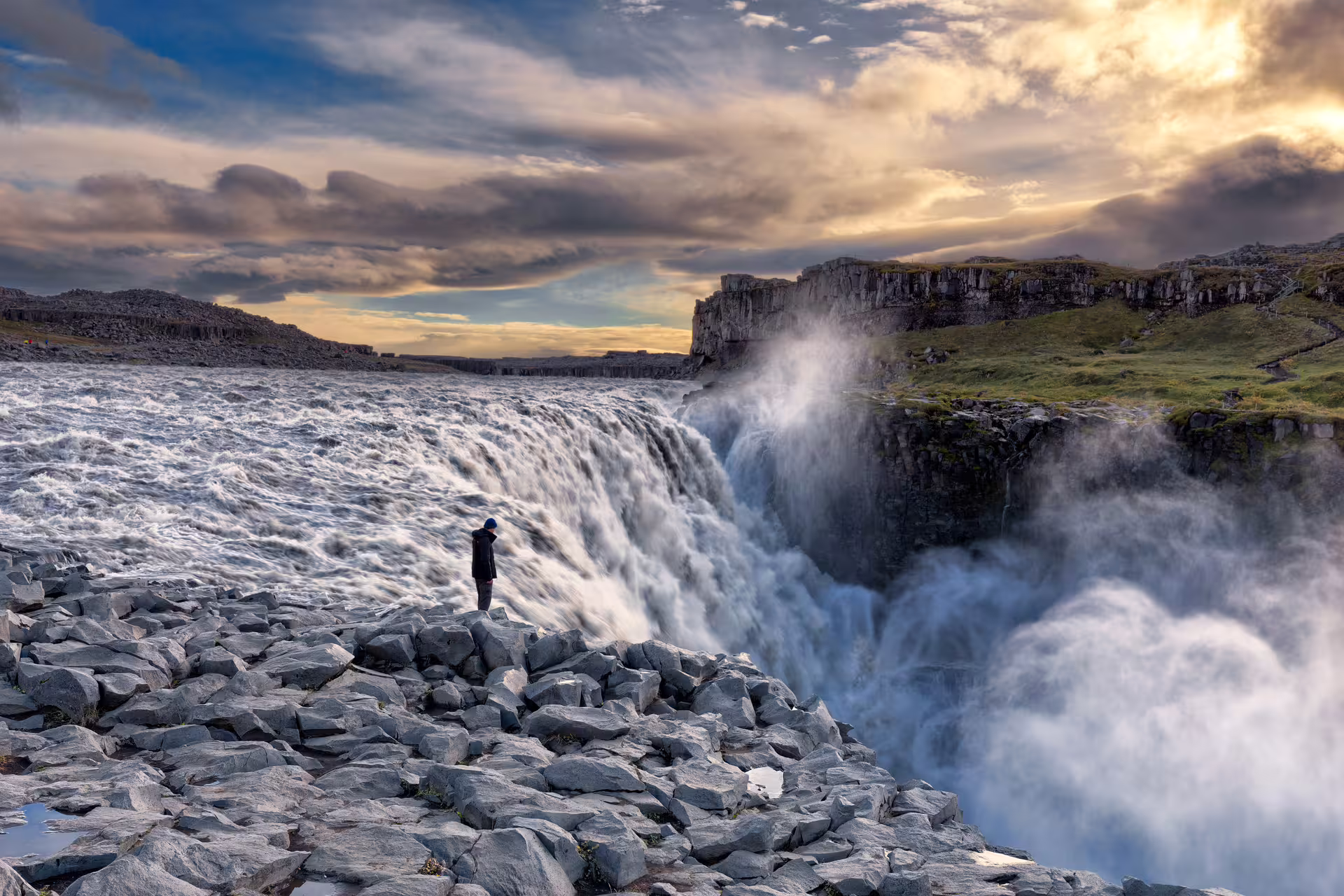 Person standing on rocky edge of Dettifoss waterfall in Iceland under dramatic cloudy sky