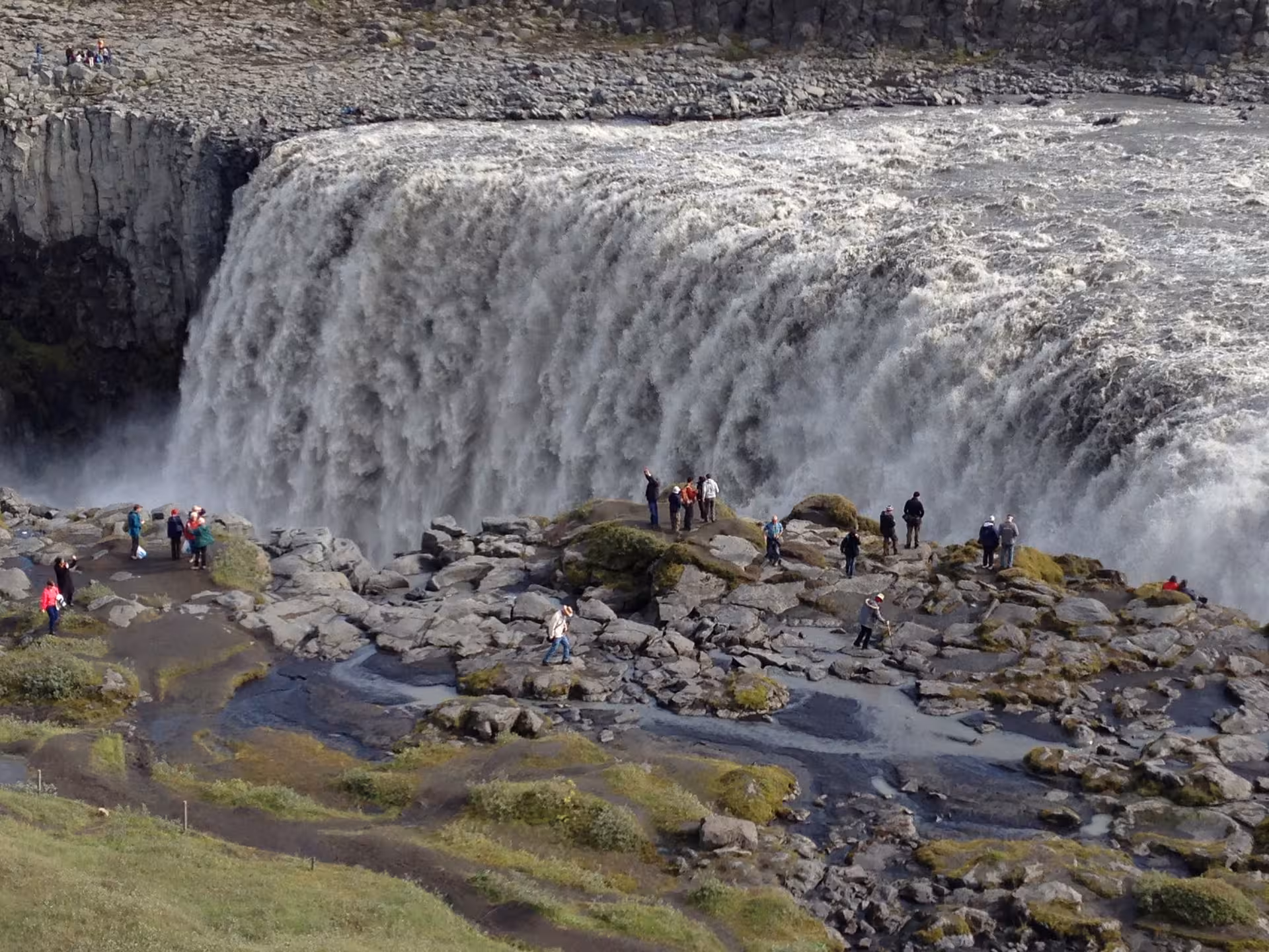 Tourists admire the powerful cascade of Dettifoss Waterfall in Iceland, surrounded by rugged, mossy terrain.