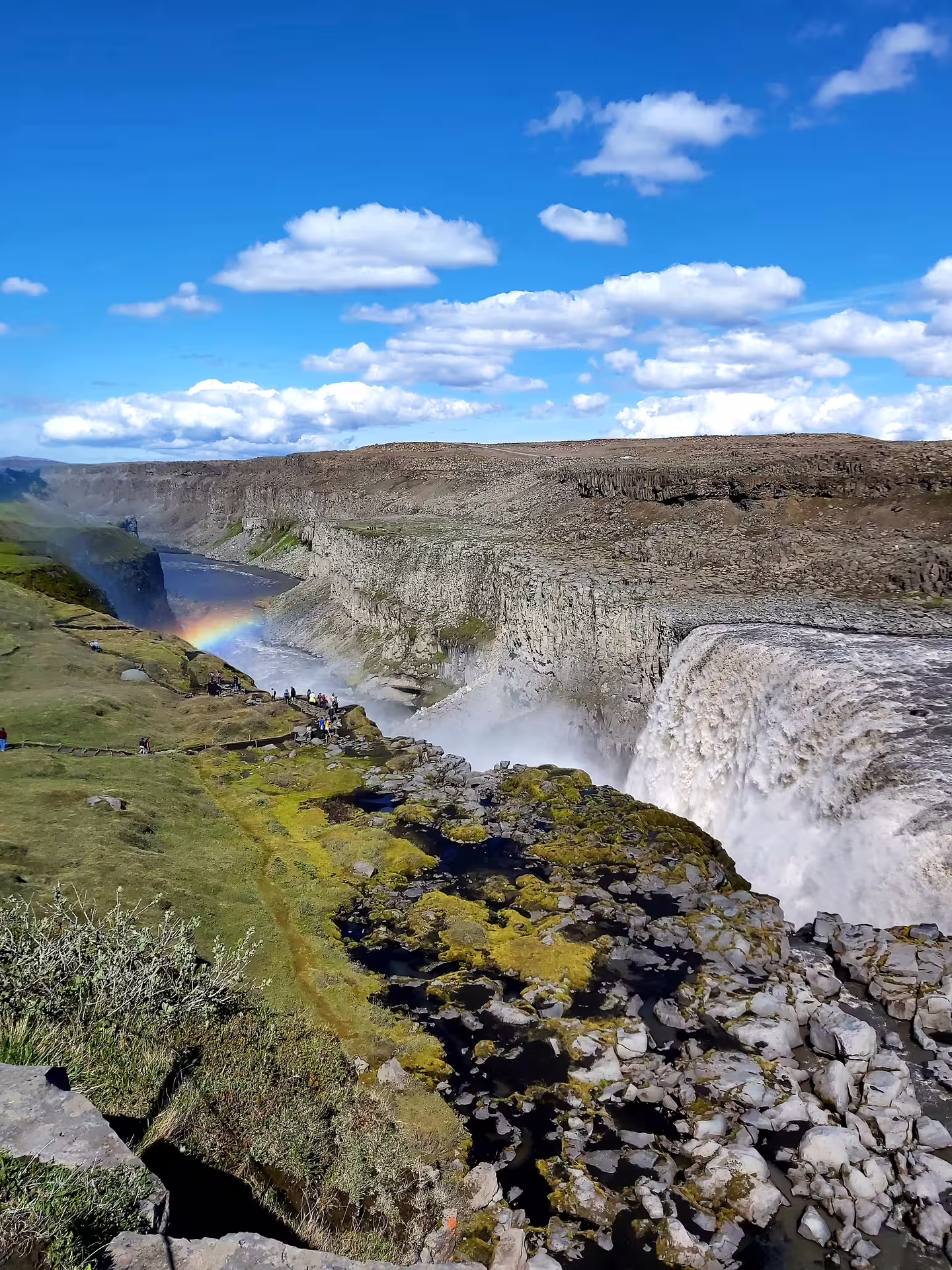 Dettifoss waterfall in Iceland with rainbow over canyon gorge and visitors on rocky cliffs under blue sky