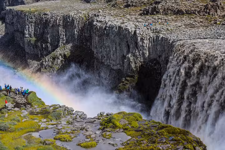 Tourists admire a vibrant rainbow over the roaring Dettifoss waterfall surrounded by lush greenery.