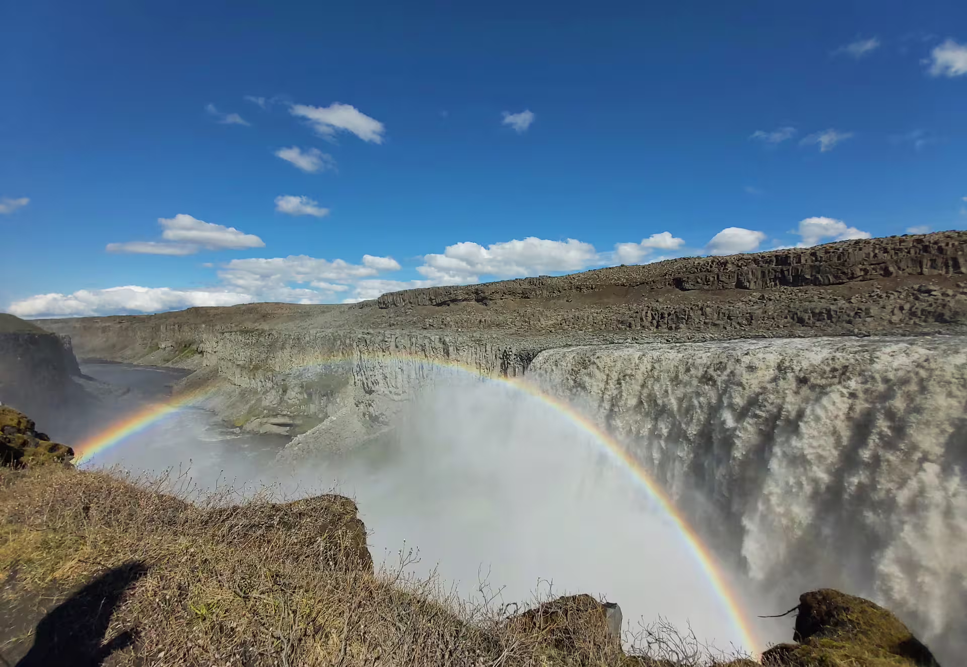 Rainbow over Dettifoss waterfall canyon with mist rising under blue sky in Iceland