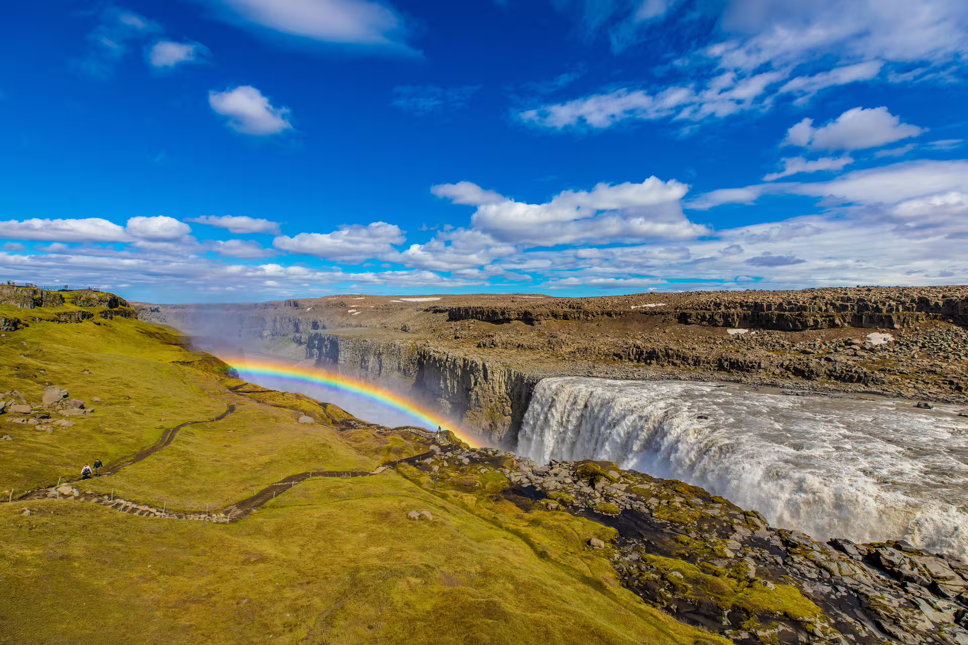 Dettifoss waterfall with rainbow over canyon, green mossy cliffs and blue sky, Iceland