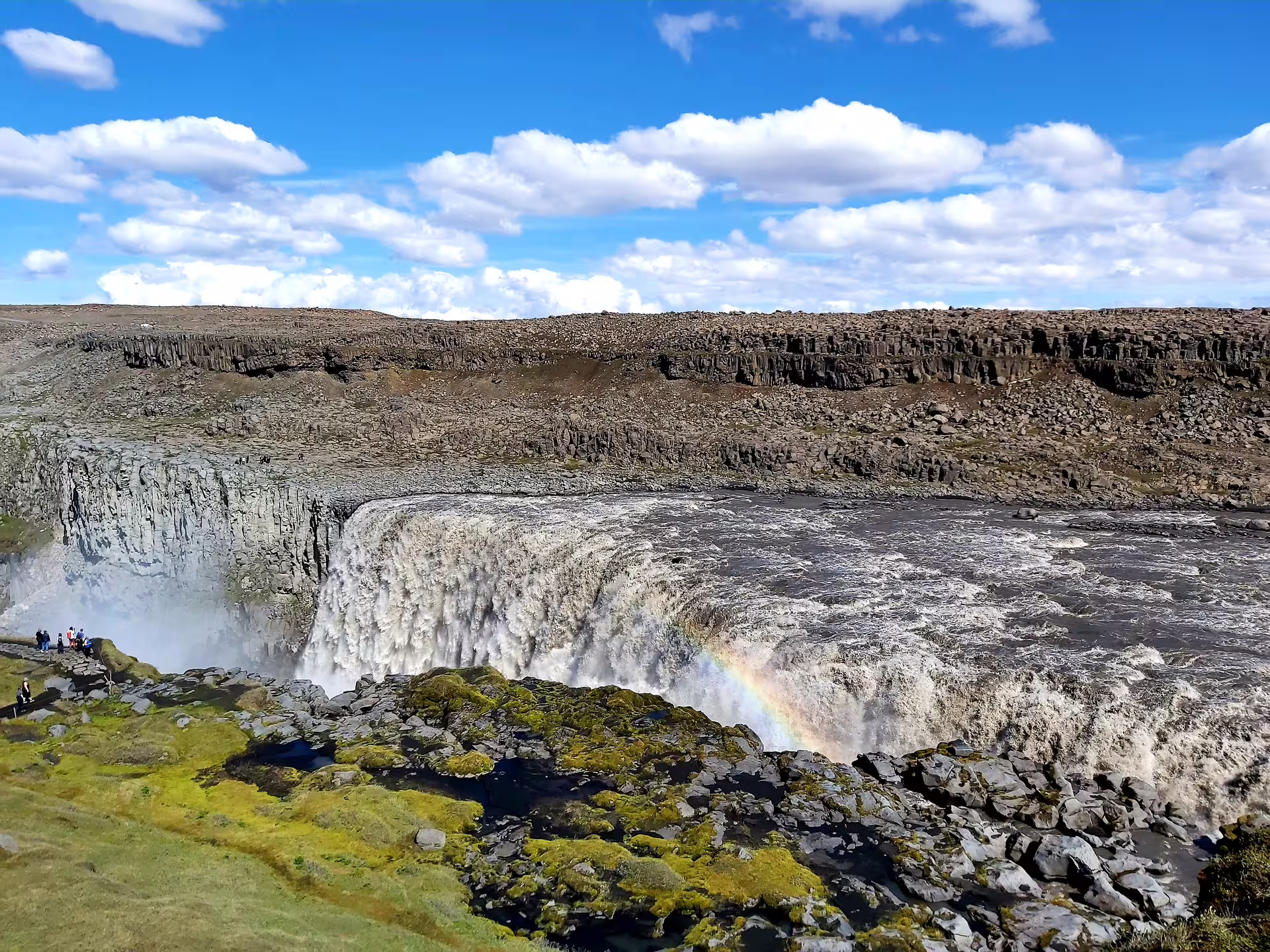 Dettifoss waterfall in Iceland with rainbow mist, basalt canyon cliffs, and tourists on the viewing platform