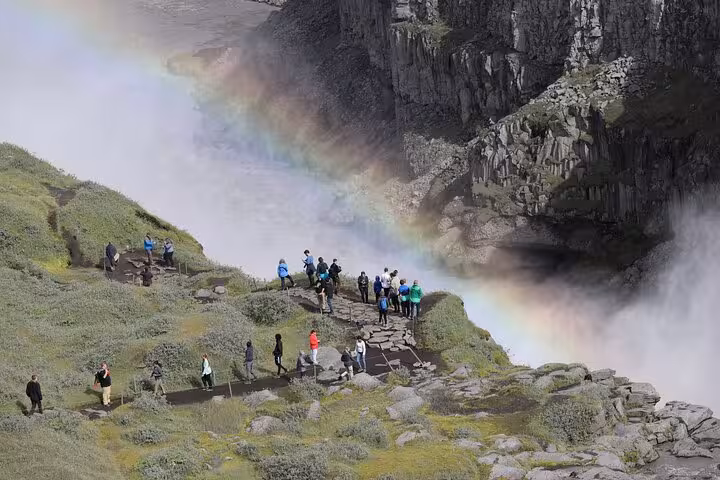 Tourists admire a vibrant rainbow over Dettifoss waterfall, surrounded by lush greenery and rocky cliffs in Iceland.