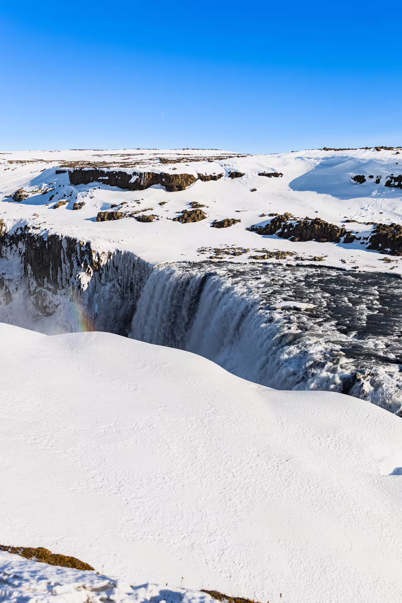 Dettifoss waterfall surrounded by snow-covered landscape with a rainbow in winter, North Iceland