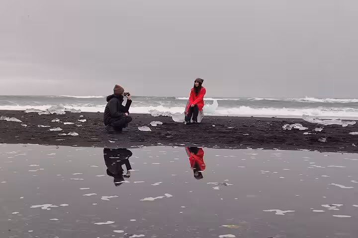 Photographer capturing woman in red jacket posing on Diamond Beach black sand with ice chunks and waves