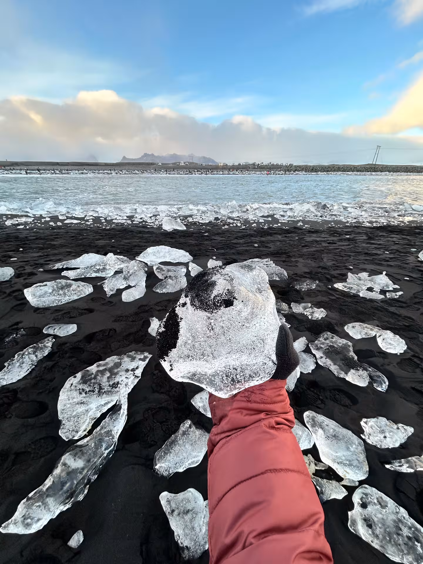 Hand holding glacial ice chunk on Diamond Beach black sand with ice fragments and ocean waves in Iceland