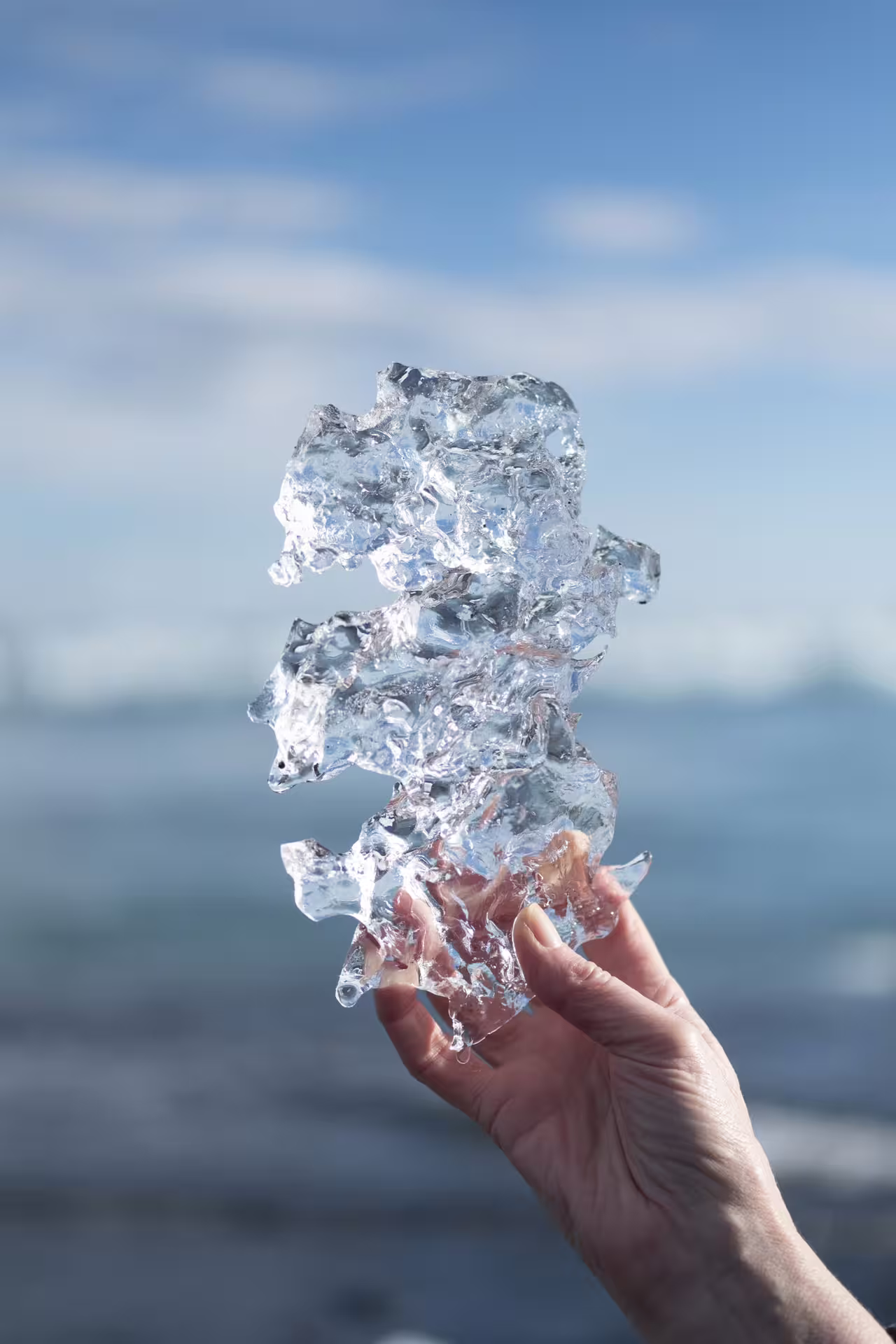 Hand holding a jagged piece of glacial ice against a blue sky and ocean backdrop at Diamond Beach Iceland