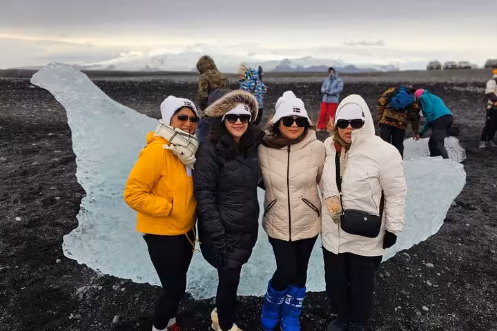 Four women in winter jackets posing near glacier ice chunk on black sand Diamond Beach Iceland