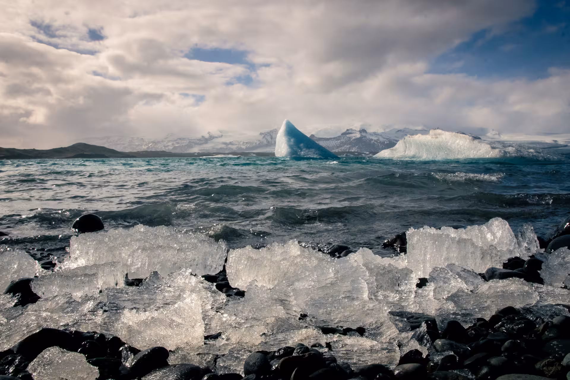 Ice chunks on black pebble beach with icebergs floating in glacier lagoon, snow-capped mountains behind
