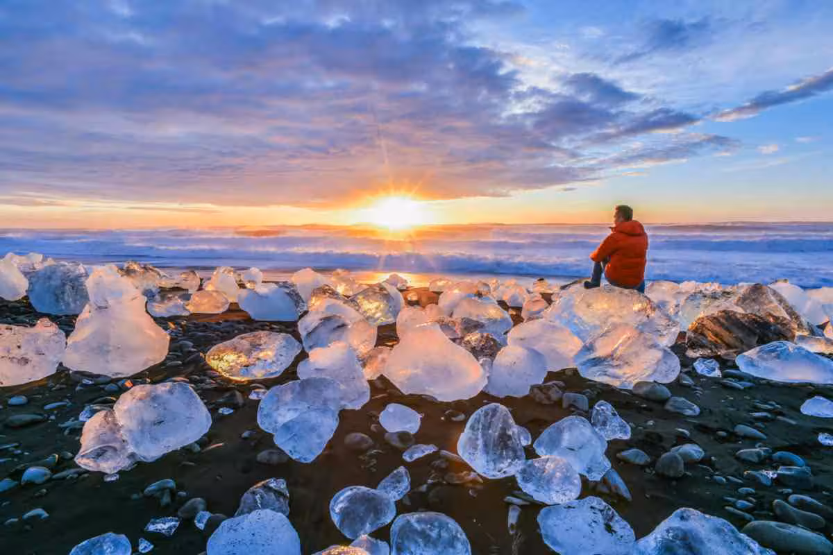 Traveler sitting on glacier ice chunks at Diamond Beach Iceland watching a stunning sunset on a South Coast private day trip from Reykjavik
