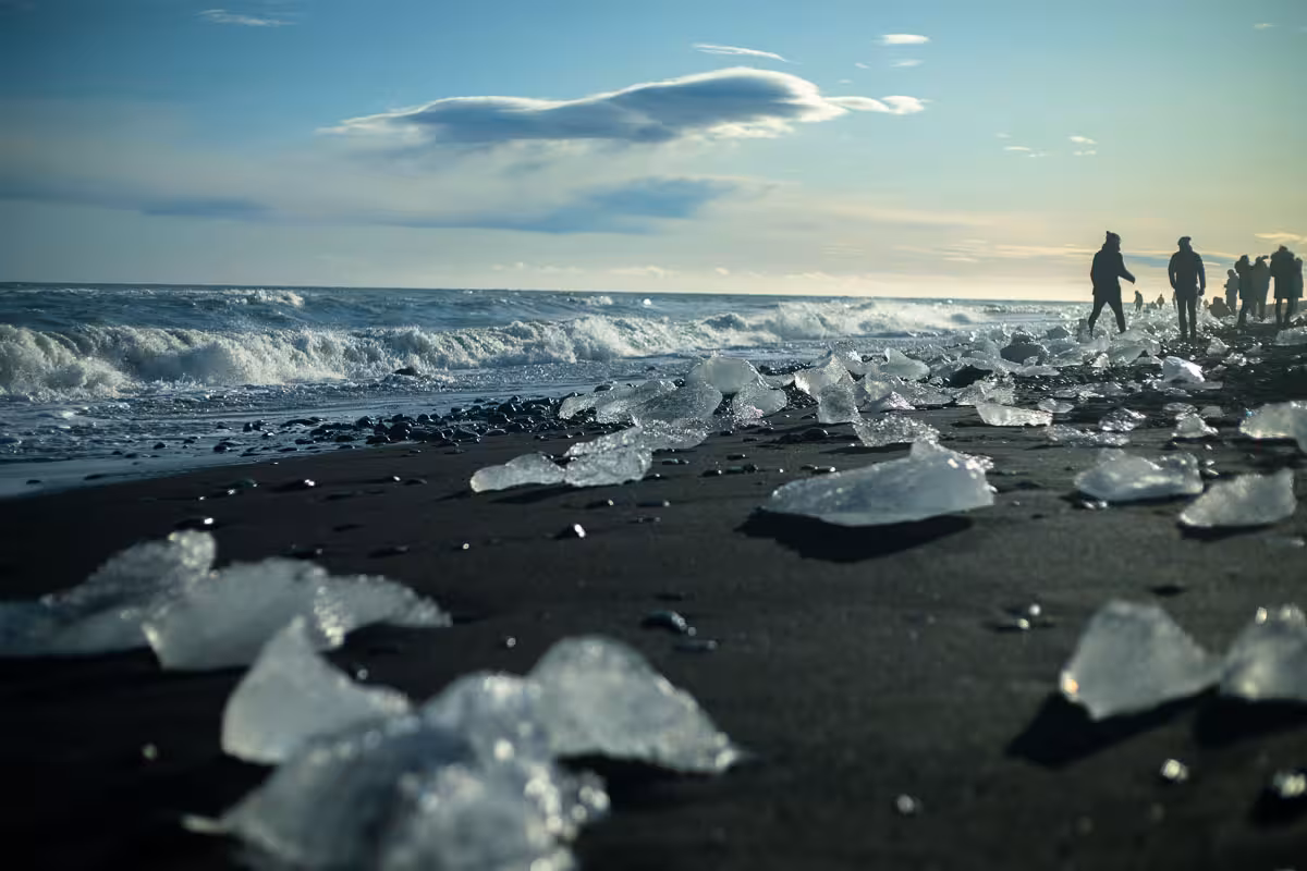 Ice chunks scattered on black sand Diamond Beach with waves and silhouetted visitors in background, Iceland