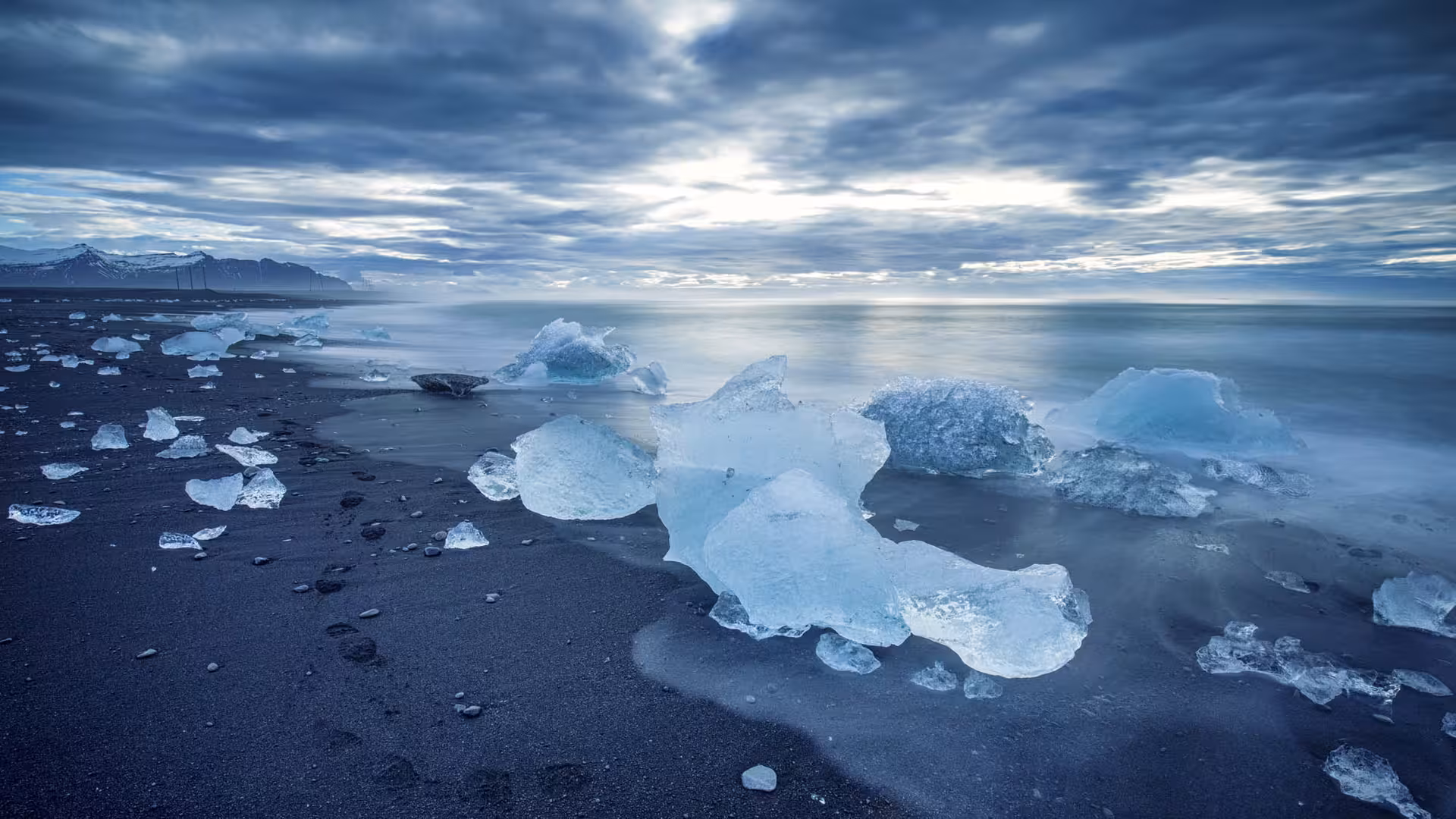 Ice chunks on black sand Diamond Beach with glacial lagoon and dramatic cloudy sky, Iceland South Coast