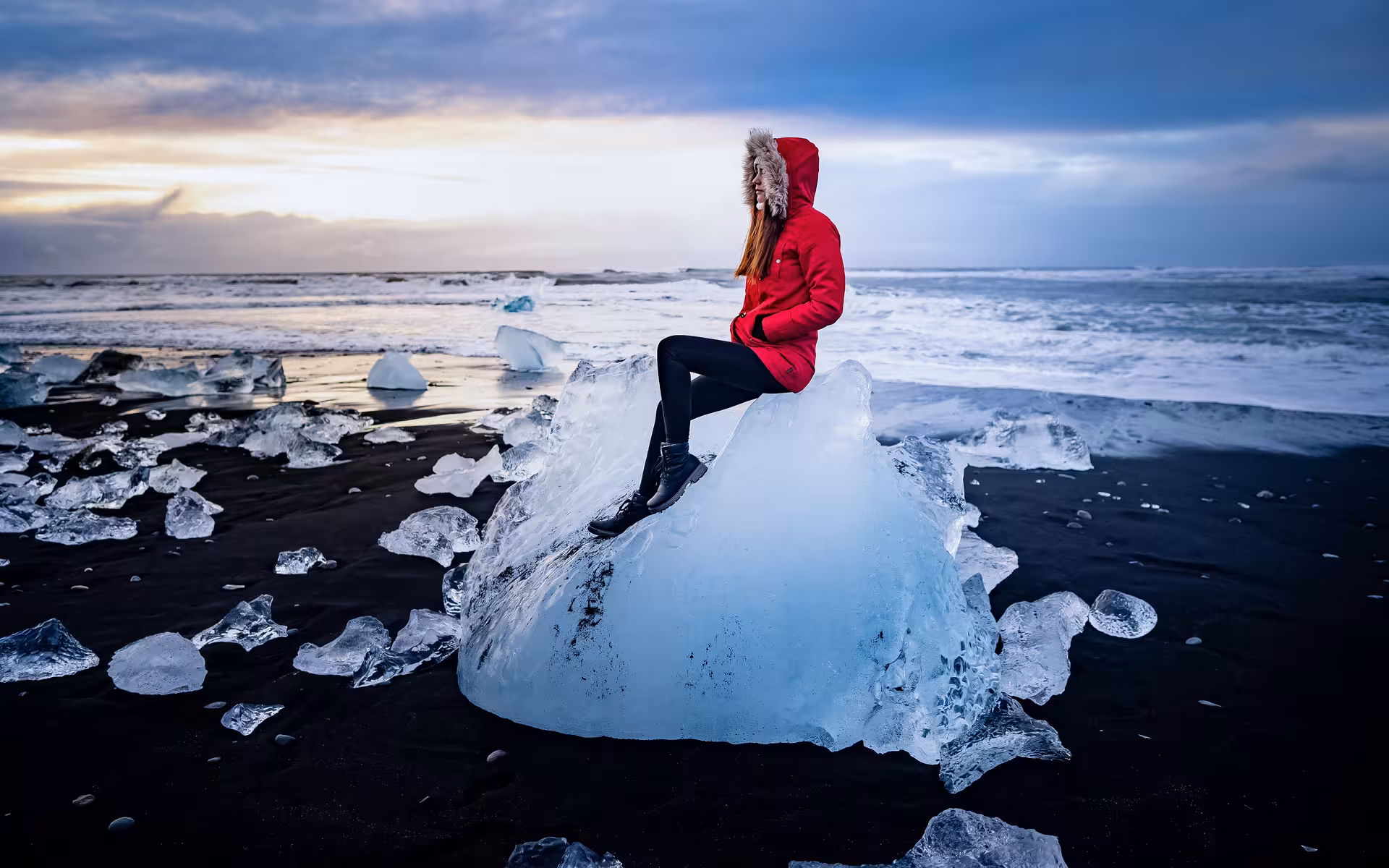 Woman in red jacket sitting on glacier ice chunk at Iceland's black sand Diamond Beach