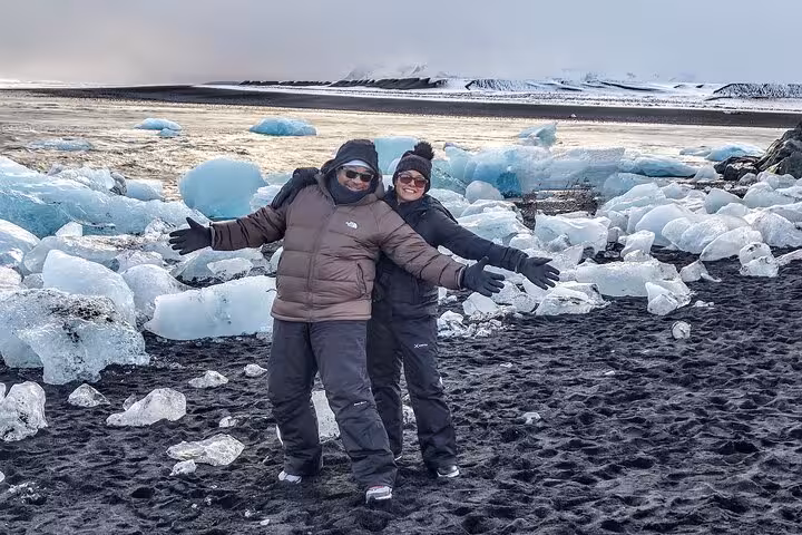 Two tourists posing on Diamond Beach black sand with blue ice chunks and glacier in Iceland