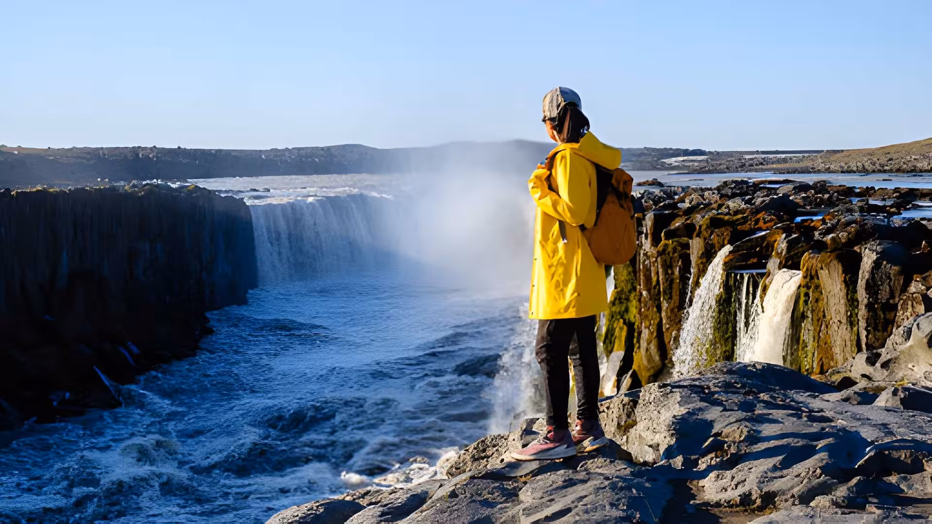 Traveler in yellow raincoat standing on basalt rocks overlooking Selfoss waterfall in Iceland