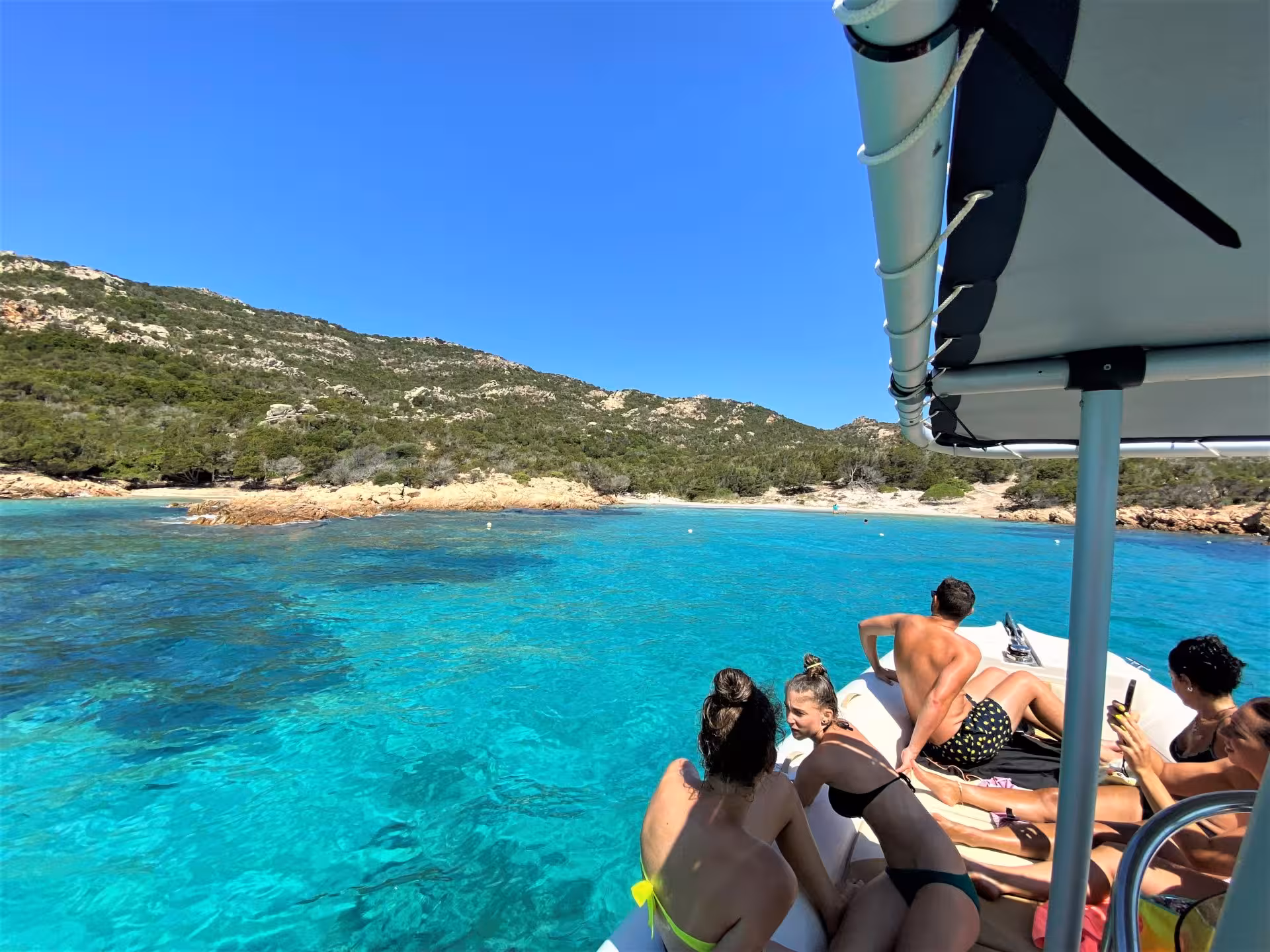 Tourists enjoy a dinghy ride on crystal-clear waters near La Maddalena's scenic beaches, surrounded by lush hills.
