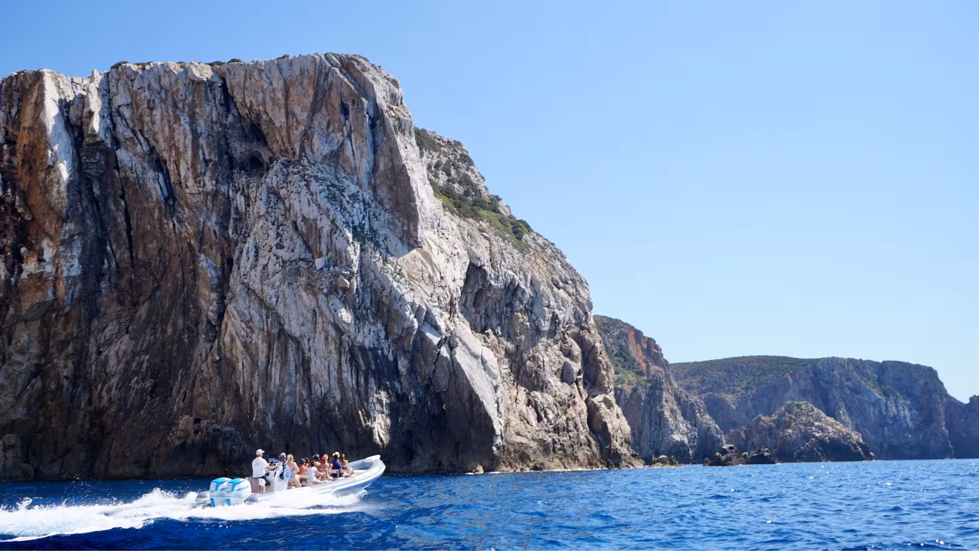 Dinghy with tourists cruising past towering cliffs near Porto Flavia on a sunny day, showcasing Sardinia's stunning landscapes.
