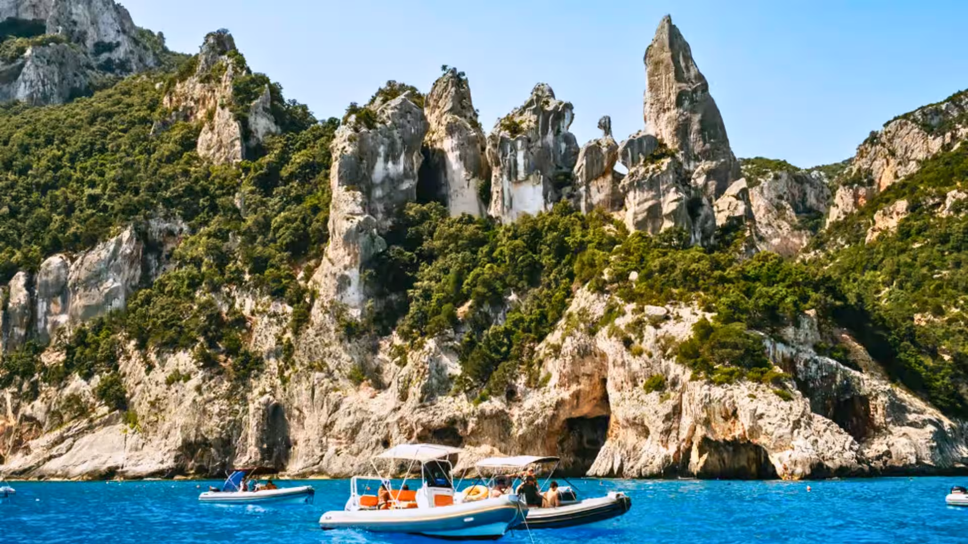 Dinghies float on the vibrant blue waters with lush cliffs in the Gulf of Orosei, Cala Gonone.