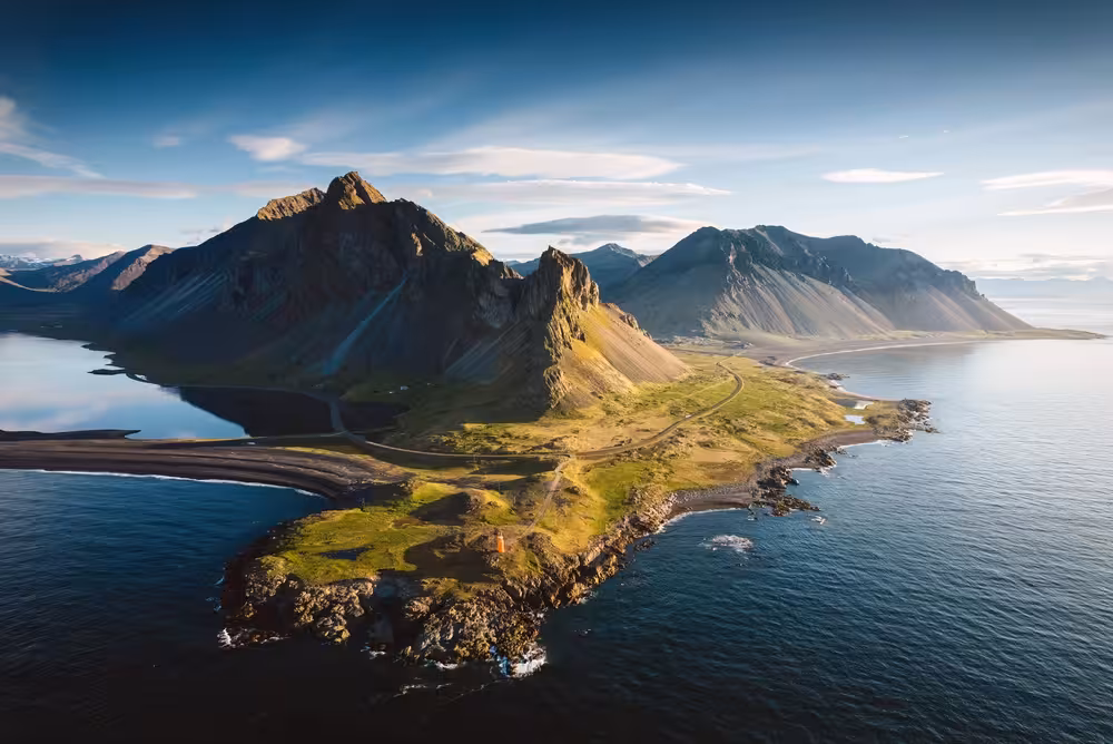 Aerial view of Icelandic coastal peninsula with dramatic black mountains, green cliffs, and calm ocean waters near Djupivogur