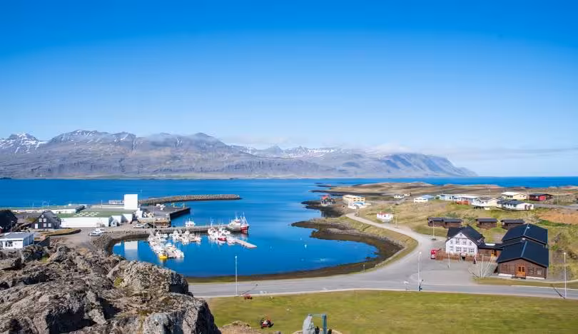 Aerial view of Djupivogur harbor village with colorful boats, calm blue fjord, and snow-capped mountains in Iceland
