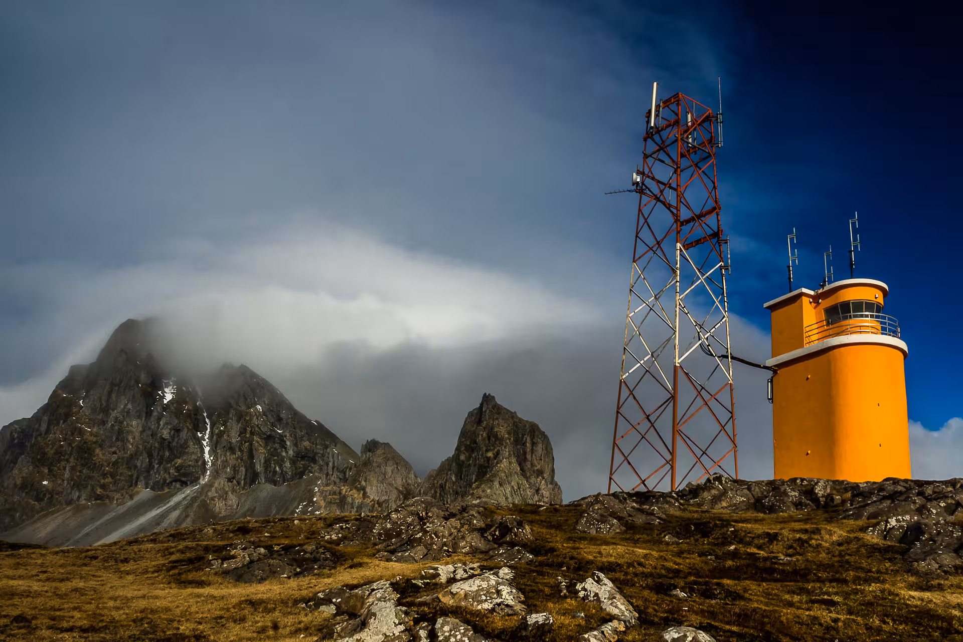 Orange lighthouse tower and red communication mast on rocky highland with dramatic misty mountains in Iceland