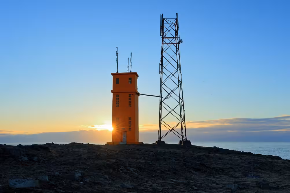 Orange lighthouse tower and communication mast at sunset on rocky Icelandic coastline